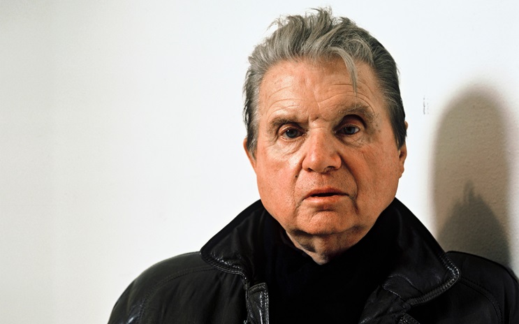 Francis Bacon wearing a black jacket stands against a plain white wall.